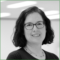 A black and white head shot of Amy Greenberg, standing inside an office space. She wears glasses and is smiling at the camera