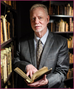 Larry P. Alford standing in front of book shelves while holding a book open in his hands.